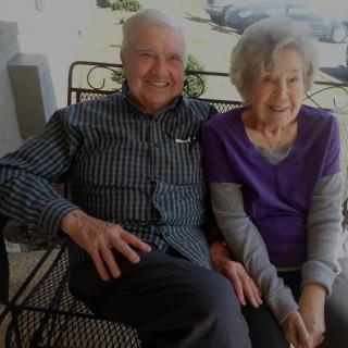 An elderly couple sitting together on a bench, smiling outdoors.