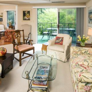 A cozy living room with a mix of traditional and modern furniture, featuring a glass coffee table and a view of the balcony through sliding glass doors.