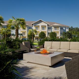 A cozy outdoor seating area with a fire pit in front of a modern residential building lined with palm trees.