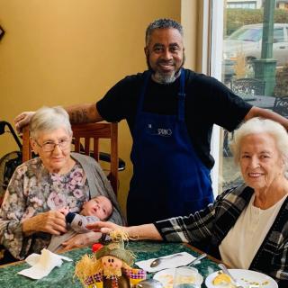 A joyful family gathering at a restaurant, featuring two seniors and an infant, with a friendly staff member.