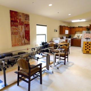 A rehabilitation room equipped with therapy exercise machines, chairs, and an adjacent kitchen area.