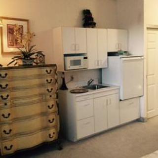 A compact kitchenette with white cabinets, a microwave, and a sink beside a decorative chest of drawers.