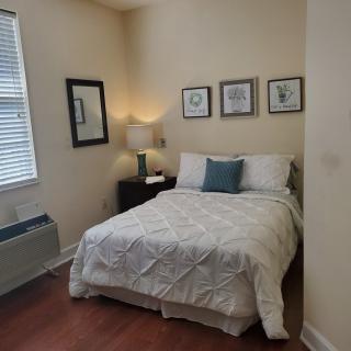 A cozy bedroom with a neatly made bed, decorative pillows, framed artwork on the wall, and a lamp on the bedside table.