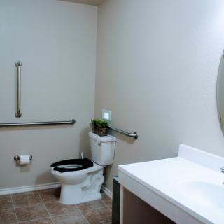A clean, handicap-accessible bathroom with a toilet, sink, mirror, and grab bars.
