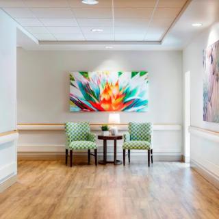 A bright and inviting hallway features two green chairs and a small table, highlighted by a colorful abstract painting on the wall.