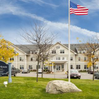 A large building with a flagpole flying the American flag, set amidst a green lawn and surrounded by parked cars under a cloudy sky.
