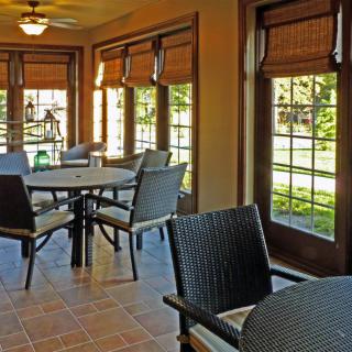 A cozy sunroom dining area with multiple tables and chairs, featuring large windows and blinds.