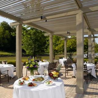A scenic outdoor dining area with elegantly set tables under a pergola, surrounded by lush greenery.