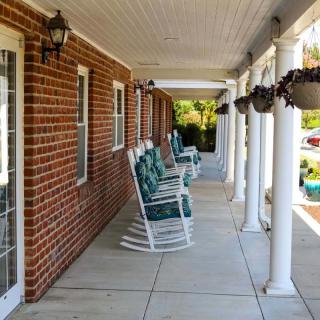 A covered porch with rocking chairs lined along a brick wall, featuring columns and hanging plants.