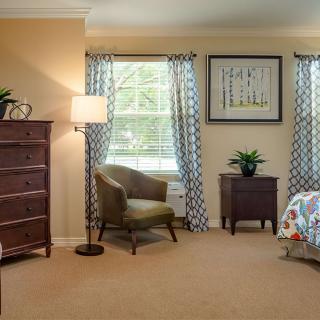 A cozy bedroom featuring a large dresser, an armchair by the window, and a neatly made bed with colorful bedding. Elegant curtains frame the windows, adding warmth to the room's decor.