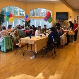A group of people enjoying a festive gathering with balloons and dining tables in a bright indoor setting.