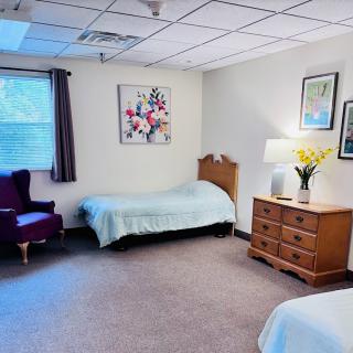 A well-lit bedroom with two single beds, a dresser with a lamp and flowers, and a comfortable chair near a window.
