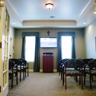 A small chapel interior with rows of chairs, a podium, and a cross on the wall.
