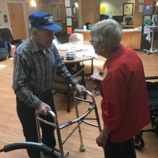 Two elderly individuals conversing indoors, one using a walker and the other in a wheelchair.