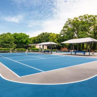 An outdoor tennis court with a blue surface, surrounded by trees and shaded seating areas.