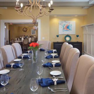 A beautifully set dining room with elegant decor, featuring a long table adorned with blue napkins and a centerpiece of red flowers under a grand chandelier.