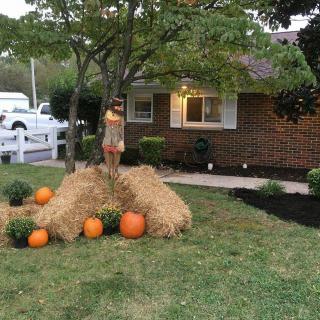 A cozy autumn-themed yard display featuring pumpkins and hay bales under a tree in front of a house.