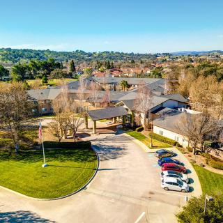 An aerial view of a building complex with a parking lot, surrounded by trees on a sunny day.
