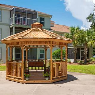 A wooden gazebo situated in front of a residential building, surrounded by green grass and trees.