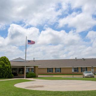 A single-story brick building with an American flag flying, surrounded by a grassy lawn and a parked car under a cloudy sky.