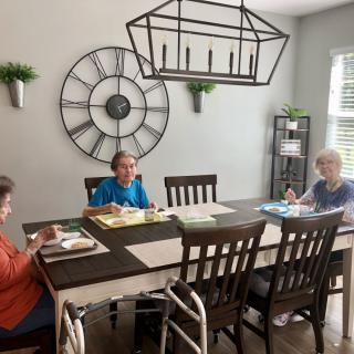 Three elderly individuals engage in an art activity around a dining table in a well-lit room.