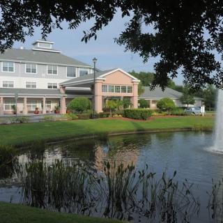 A scenic view of a building by a pond with a fountain and surrounding greenery.