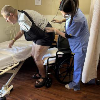 A nurse assists an elderly man transitioning from a wheelchair to a bed in a healthcare setting.