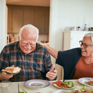 An elderly couple shares a joyful moment over a meal in their kitchen.