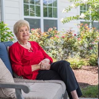 An elderly woman sitting comfortably on a patio chair outdoors, surrounded by a garden.