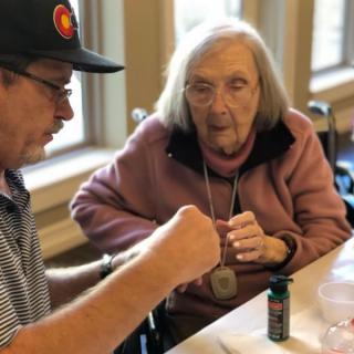 An elderly woman in a wheelchair is interacting with a man wearing a hat indoors.