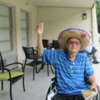 An elderly person sitting on a patio in a wheelchair, wearing a sombrero and a striped shirt, raising one hand.
