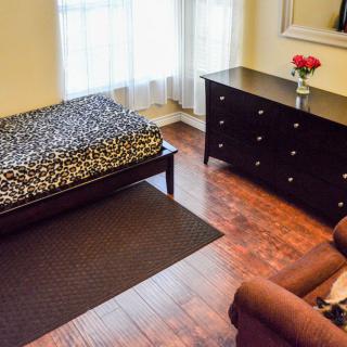 A cozy bedroom featuring a single bed with patterned bedding, a dark wooden dresser topped with a vase of roses, an armchair, and a large window letting in natural light.