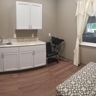 A small room featuring a sink with cabinets, a chair, a window with curtains, and part of a bed.