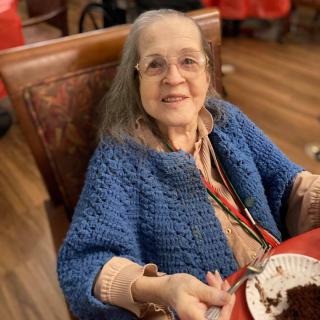 An elderly woman wearing a blue sweater smiles while enjoying dessert at a dining table.