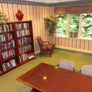 A cozy library space with a bookshelf, table, and chairs near a window with a view of greenery.