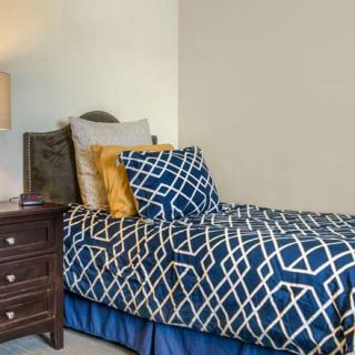 A cozy bedroom featuring a single bed with geometric patterned bedding and assorted pillows, next to a wooden nightstand with a lamp.