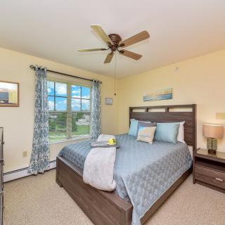 A cozy bedroom with a neatly made bed, ceiling fan, and large window with patterned curtains.