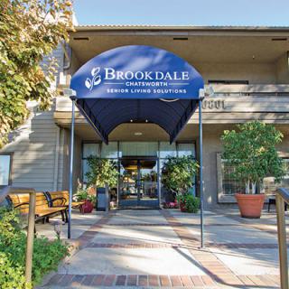 Entrance to a building with a blue canopy marked 'Brookdale', surrounded by plants and benches.