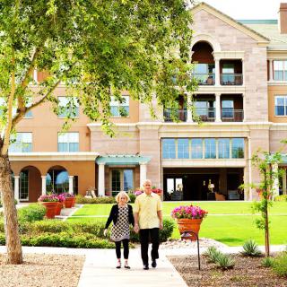 An elderly couple walking towards a retirement home surrounded by a garden.