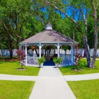 A charming white gazebo is centered in a neatly landscaped garden, surrounded by lush green grass and trees, with pathways leading up to it.