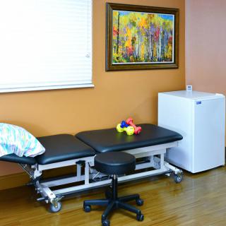 A therapy room featuring a treatment table with a pillow, a stool, a colorful toy, and a painting on the wall. There is also a small fridge in the corner of the room.