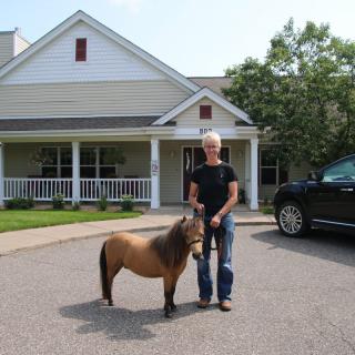 A person standing with a miniature horse in a driveway in front of a residential building.