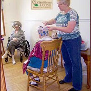 An elderly woman sits in a wheelchair while another receives a haircut in a community care setting.