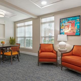 A cozy living room setting featuring orange chairs, a table with seating, artwork on the wall, and large windows that allow ample natural light.