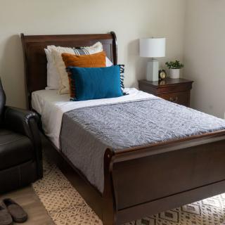 A cozy bedroom with a neatly made bed, decorative pillows, a bedside lamp, and a comfortable chair.