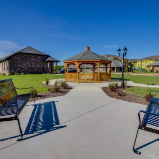 A picturesque outdoor area featuring a gazebo surrounded by benches, a well-maintained lawn, and adjacent buildings under a clear blue sky.