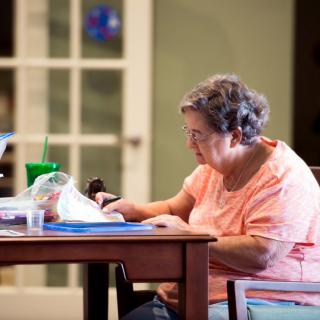 An elderly woman sitting at a table, reading a book indoors.