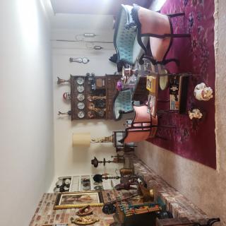 A cozy living room with vintage furniture and antique decor, featuring a red carpet and various decorative items on shelves.