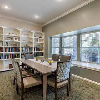 A cozy library with bookshelves, a table surrounded by chairs, and a large window letting in natural light.
