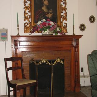 A classic indoor setting featuring a wooden fireplace mantel adorned with candles, a floral arrangement, and a portrait. A single chair rests at the side.